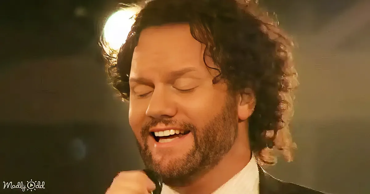 A man with curly hair and a beard is singing passionately, eyes closed, with a microphone in hand.