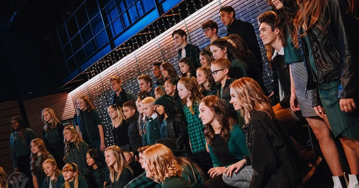 A group of children in green and black attire sing together in front of a string-lit backdrop.