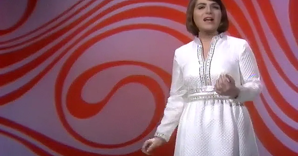 A person in a white dress with intricate patterns, smiling and gesturing with hands, against a red and white spiral...