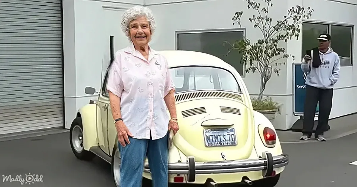 An elderly woman stands beside a vintage yellow Volkswagen Beetle, smiling in front of a white building.