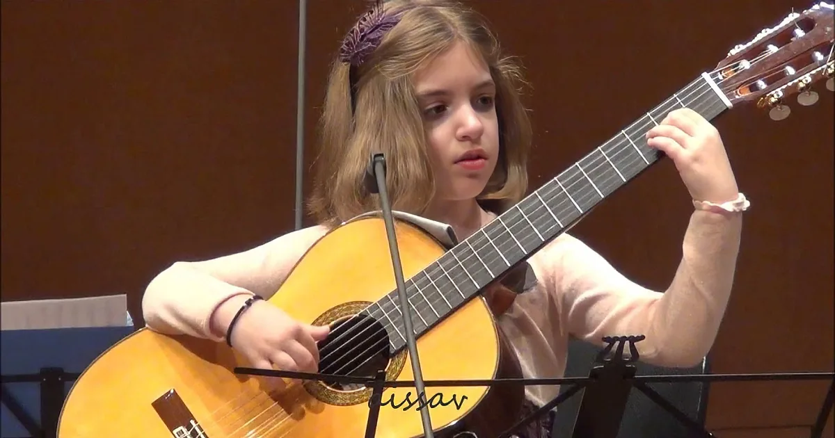 A young girl plays a classical guitar in a music setting with sheet music stands.