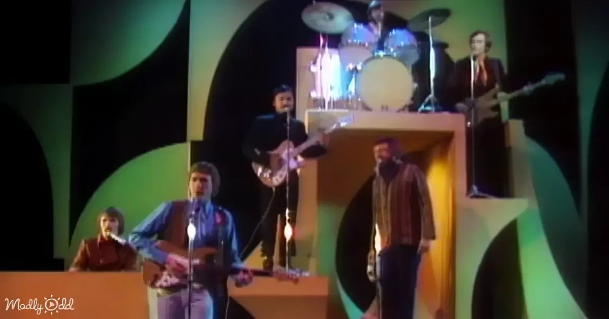 A band performs on stage with colorful geometric background, featuring guitarists, a drummer, and a lead singer.