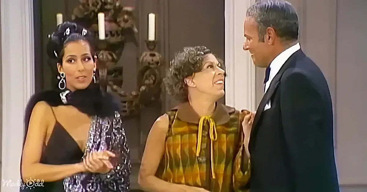 A woman in a black dress with fur collar gestures while talking to another woman and a man in a suit.