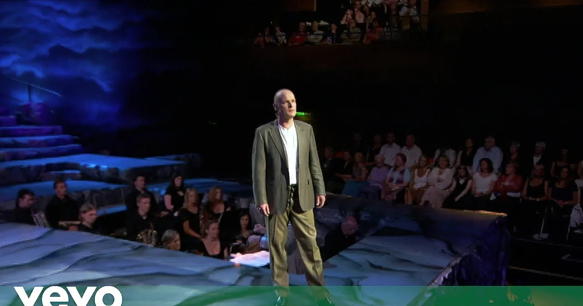 A bald man in a suit with a microphone in his ear, speaking on a stage with colorful lights.