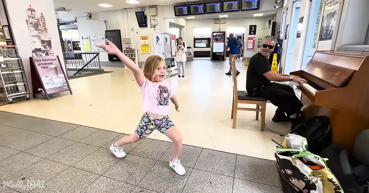 A young girl dances enthusiastically in a station, while a man plays piano nearby.