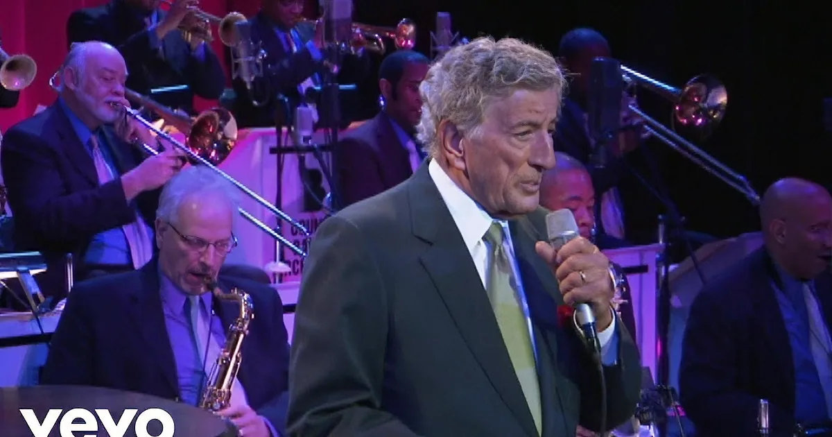 A man in a suit sings into a microphone on stage, with another person in the background.
