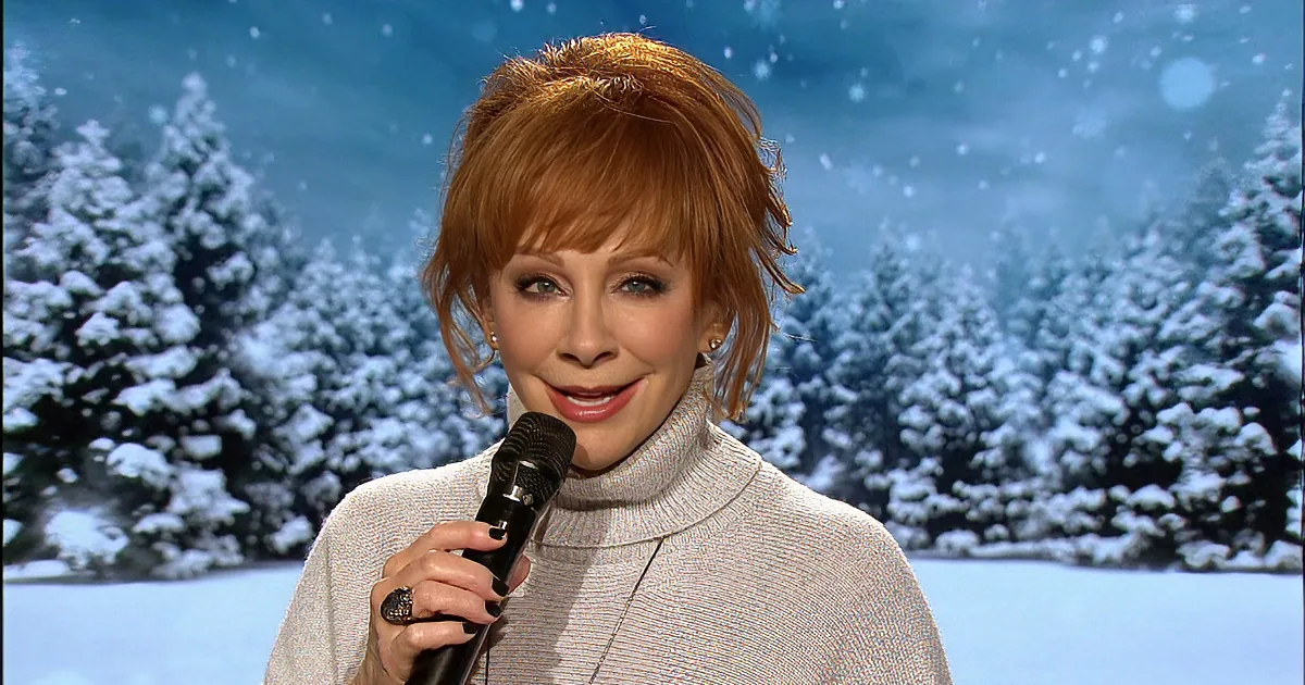 A woman with red hair and a gray sweater sings into a microphone against a snowy backdrop.