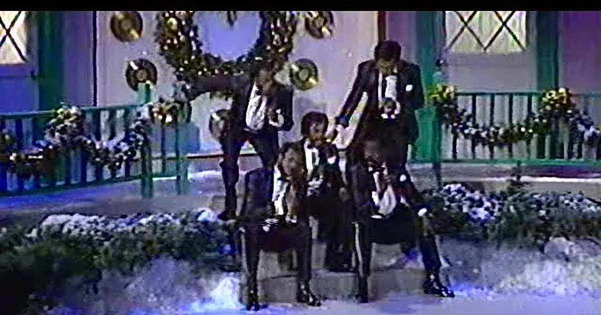 A man in a sparkly black suit sings into a microphone, eyes closed, with a golden cymbal in the background.
