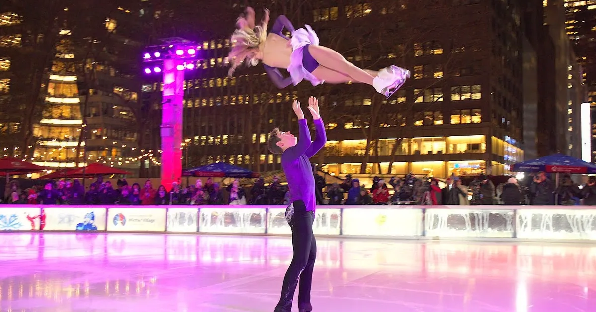 A figure skater lifts his partner in a dramatic pose on an ice rink, surrounded by spectators.