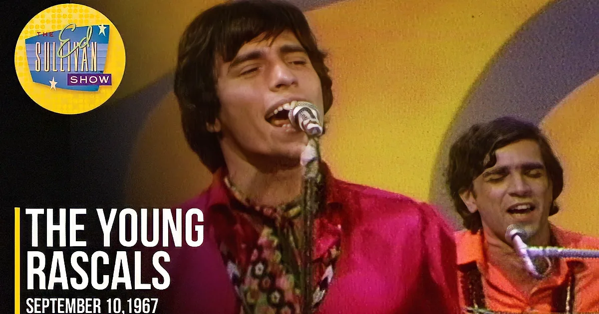 A man in an orange shirt sings into a microphone, with a drummer in the background and a large yellow and blue backdrop.