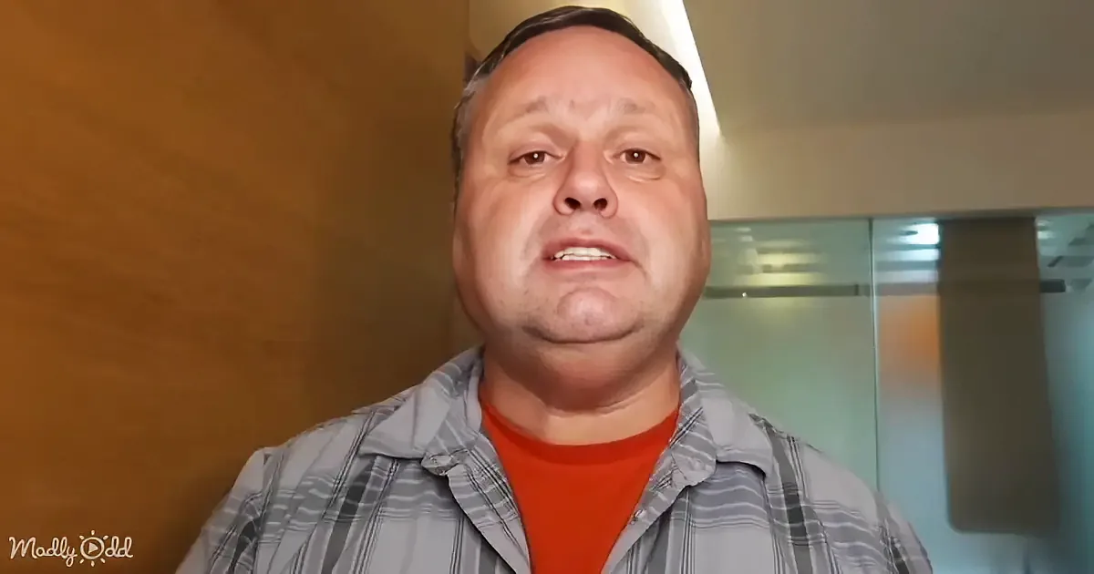 A man in a striped shirt and red undershirt looks directly at the camera with a neutral expression.