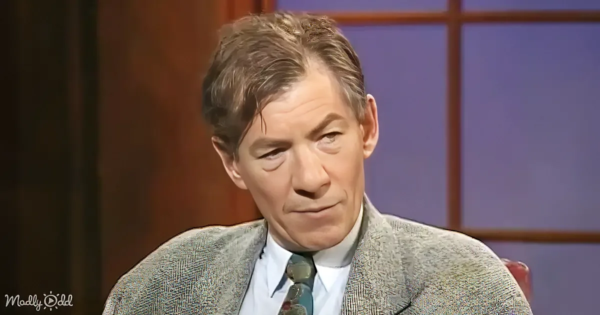 A man in a textured suit jacket and tie, seated, looking downward, with a window and wooden paneling in the background.