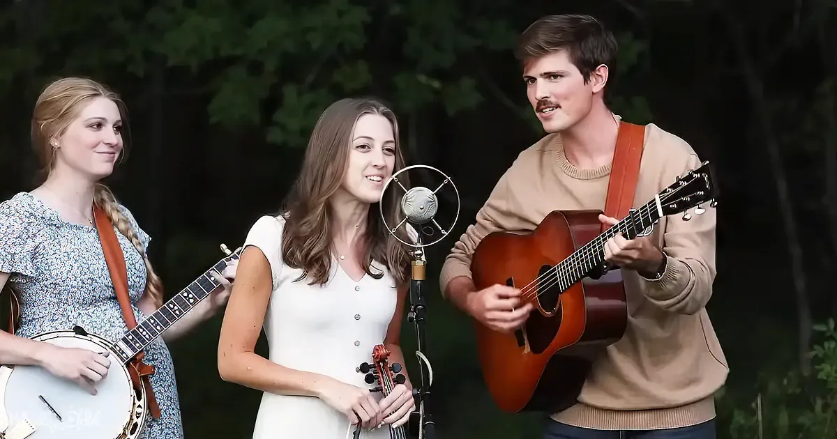 A woman in white sings into a microphone, flanked by a man playing guitar and another woman with a banjo.