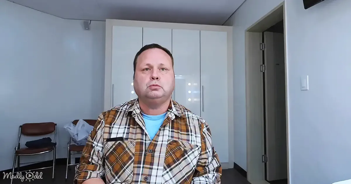 A man in a plaid shirt sits in a room with a chair and a closet in the background.