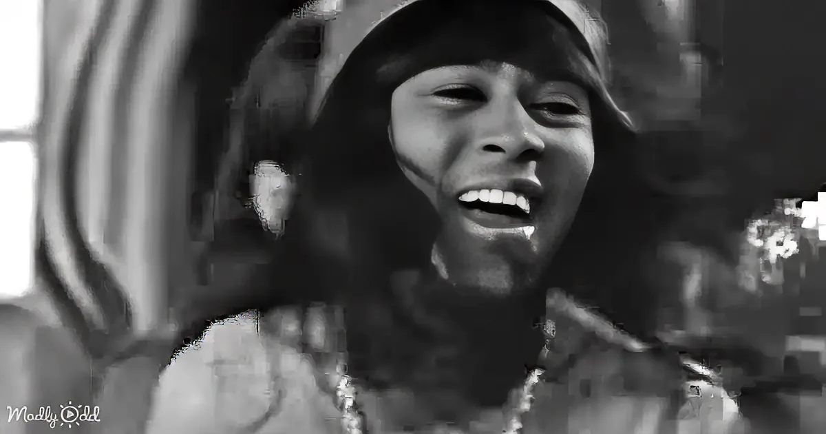 A person with long hair and a hat is smiling, wearing a patterned top, in a black-and-white image.