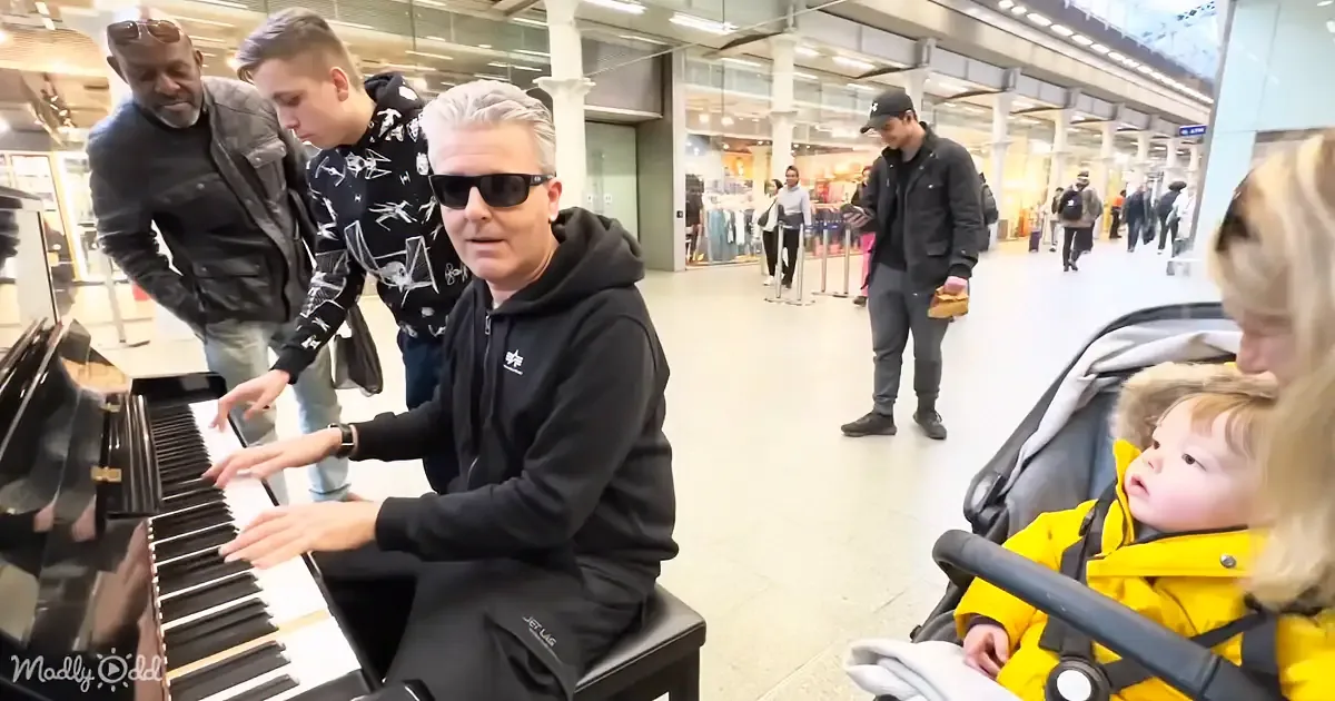 A man in a black hoodie plays a piano in a busy shopping area, surrounded by onlookers.