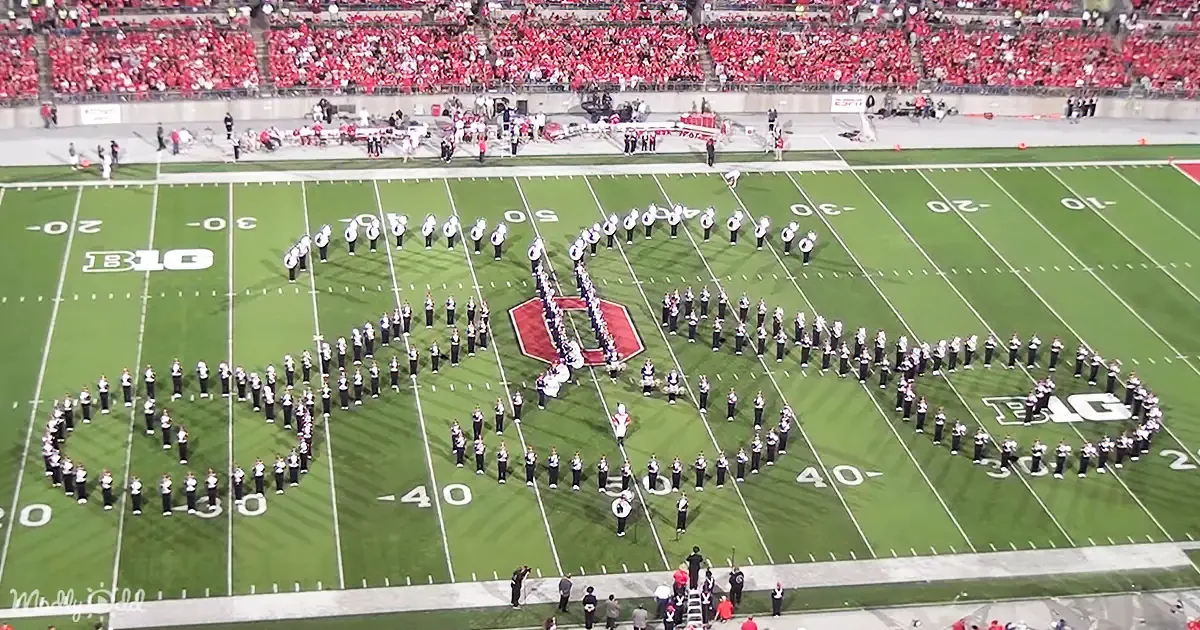 A marching band forms a large "B" on a football field with a crowd in the background.