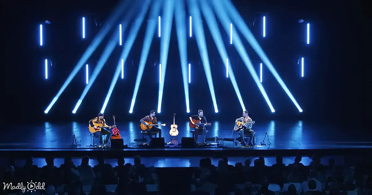 Four musicians play acoustic guitars on stage under bright blue spotlights, with an audience in the foreground.
