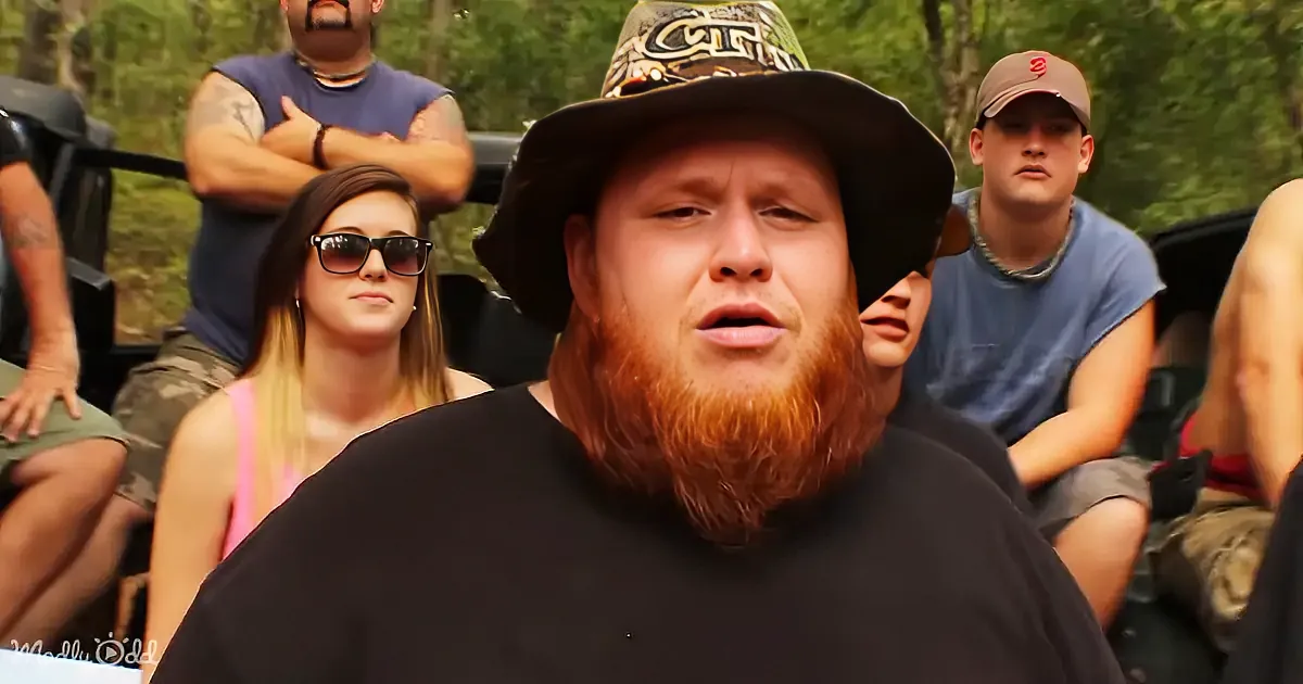 A man with a red beard and black shirt, wearing a hat, sits in front of a group of people in a vehicle, surrounded by trees.