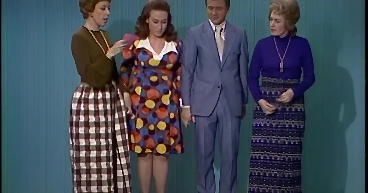 A woman in a brown top and necklace stands, while another hugs her in a colorful dress and blue suit.