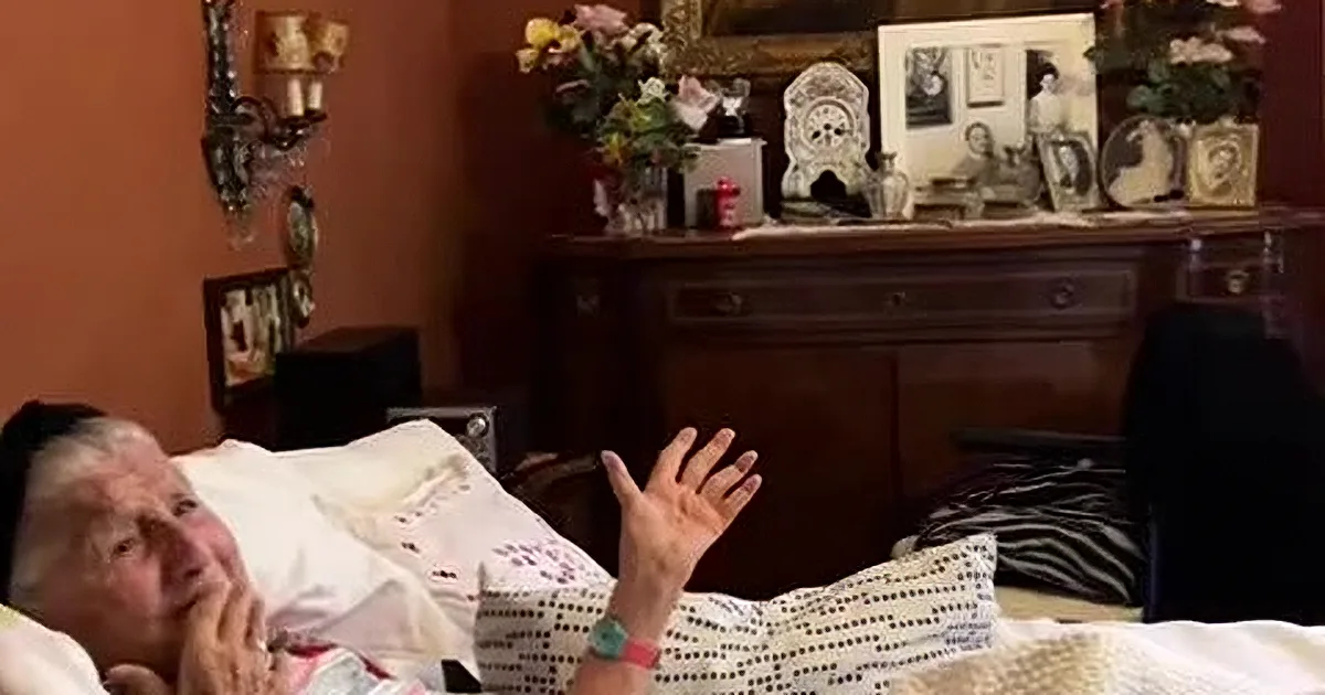 An elderly woman in a hospital bed holds a folded paper, while two men in suits play music nearby.