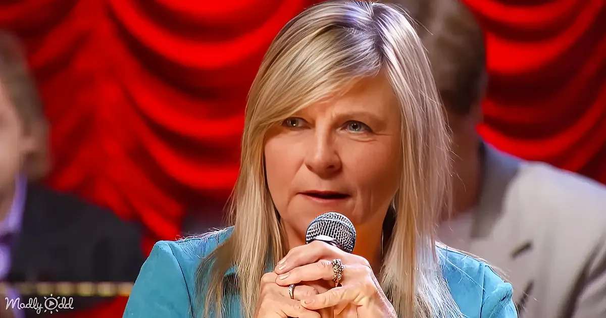 A woman with blonde hair, wearing a blue outfit, holds a microphone, with a red curtain and audience in the background.