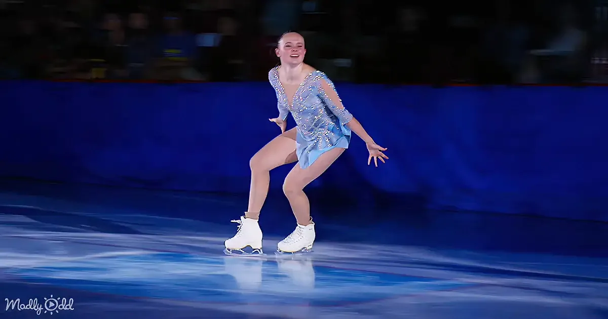 A figure skater in a blue dress glides gracefully on ice, smiling with arms outstretched.