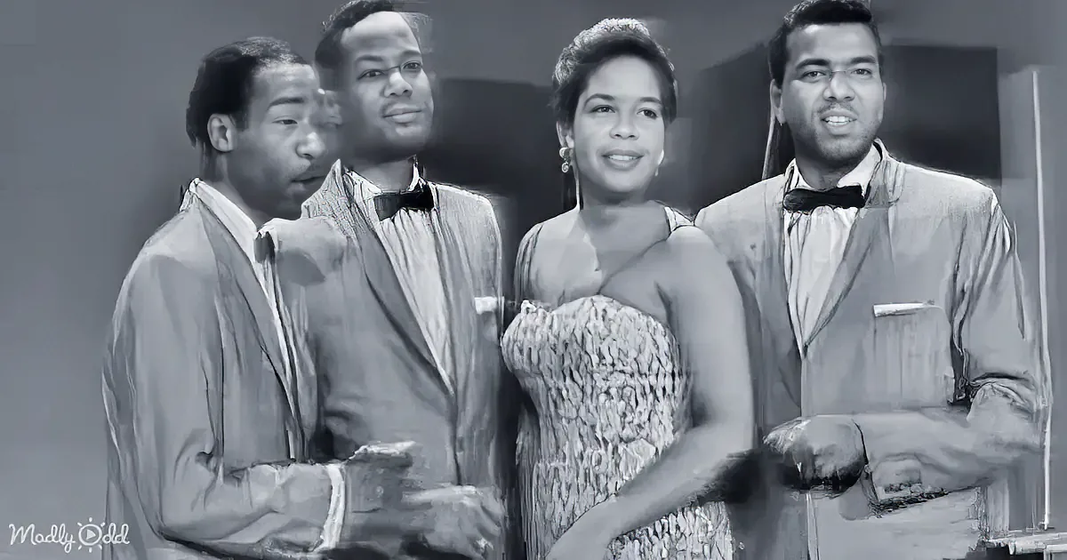 A woman in a textured dress stands with three men in formal attire, possibly from a vintage performance.