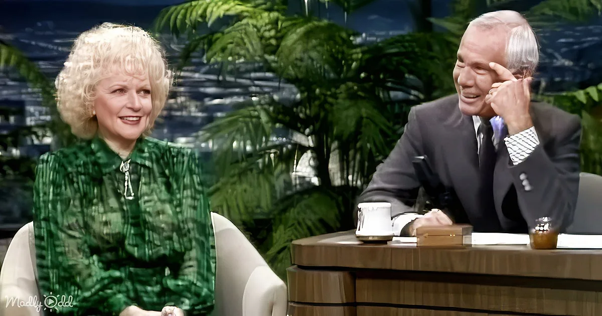 Woman in green dress smiles while seated, man in suit leans forward, both in a talk show setting with plants in the...