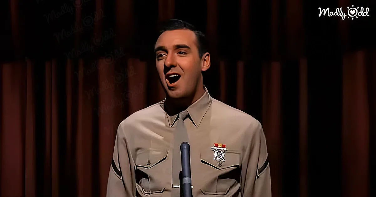 A man in a uniform sings into a microphone, wearing a badge and standing in front of a dark curtain.