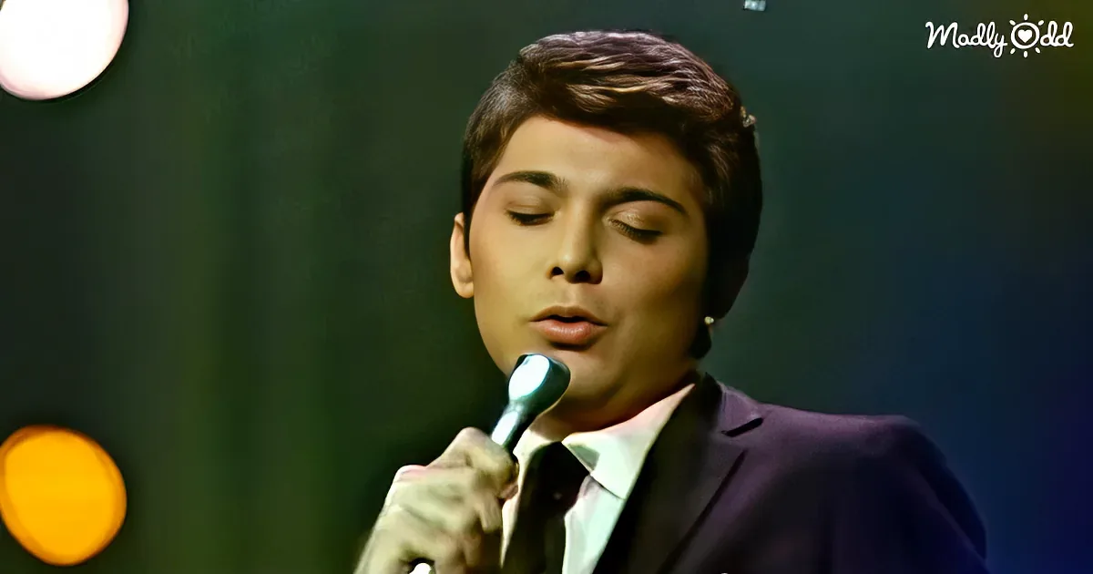 A person in a suit holds a microphone, eyes closed, against a dark background with a spotlight.