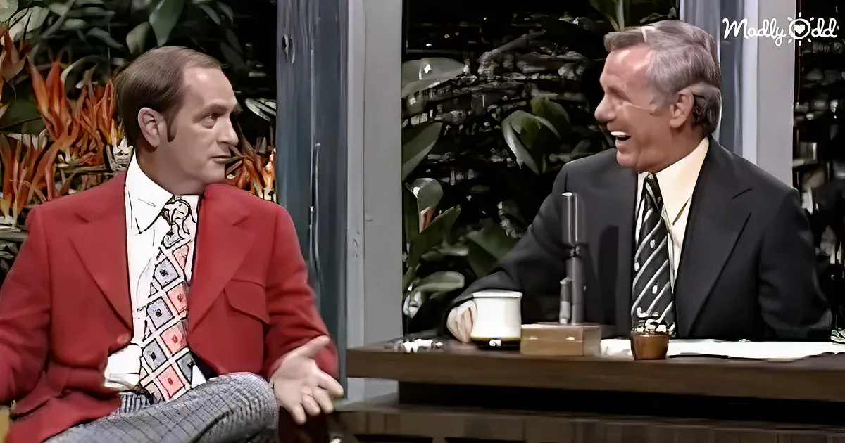 A man in a red suit gestures while seated next to another man in a suit, both in a talk show setting with plants and a...