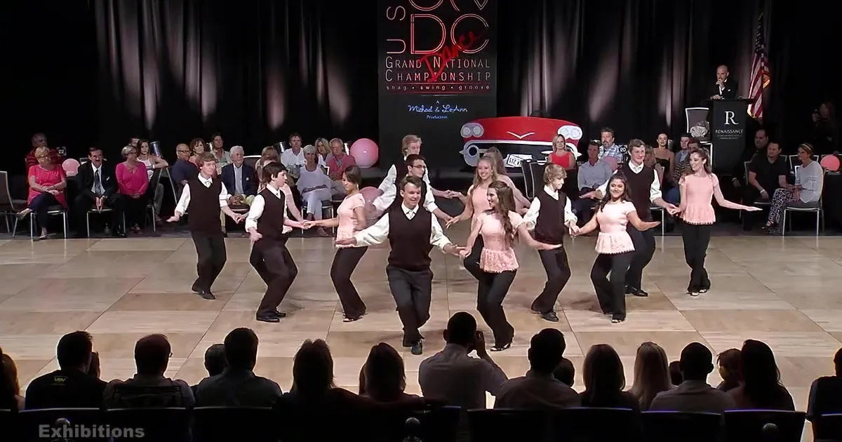 A couple dances on stage, audience watches, man in vest, woman in pink top, formal attire.