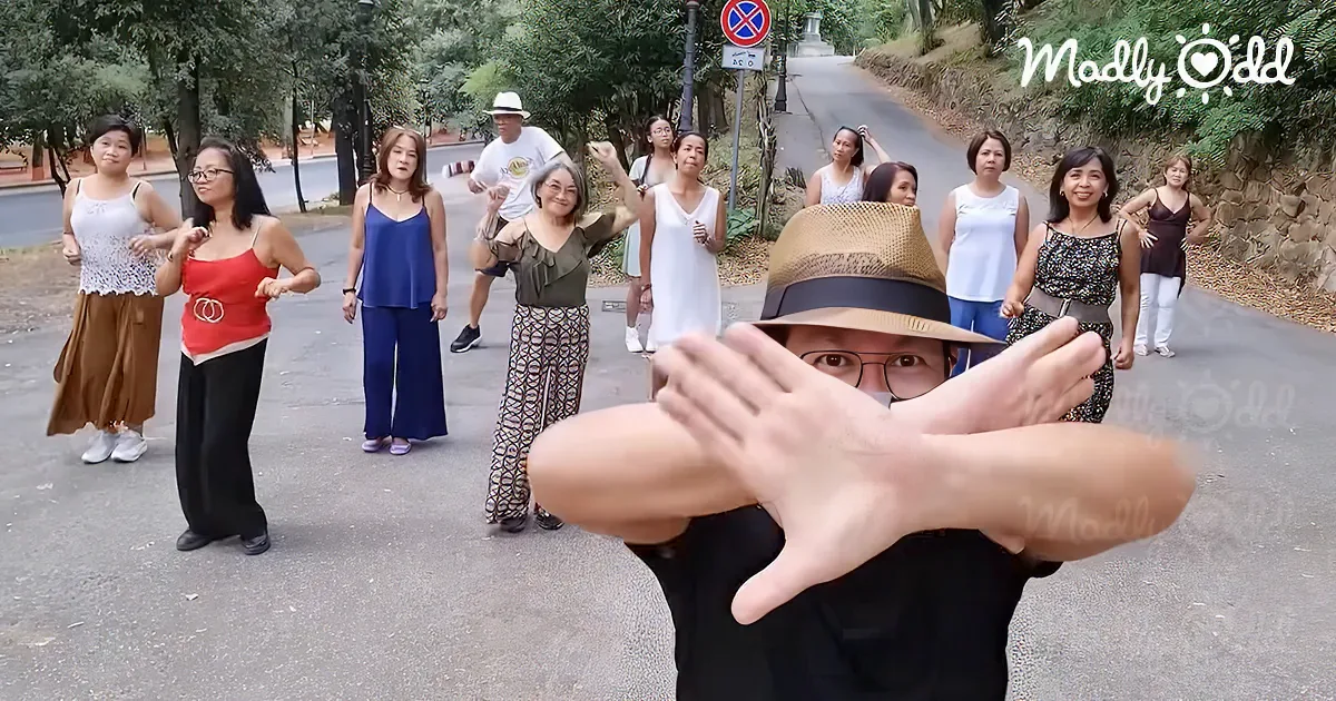 A person in a hat and glasses crosses their arms in front of their face, with a group of people dancing behind them on a...