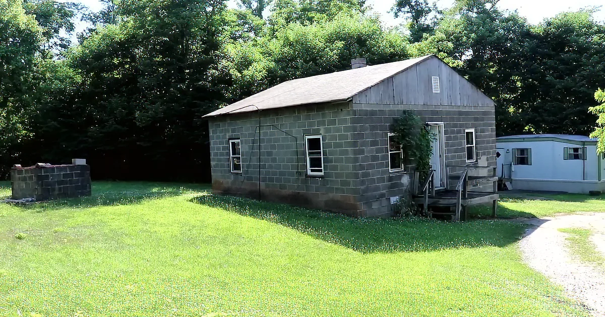 A small, gray, single-story house with a slanted roof sits on a grassy lawn surrounded by trees.