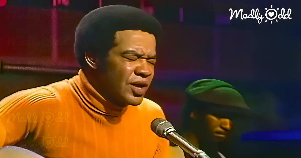 A man in an orange shirt sings into a microphone, with a blurred figure in a green mask in the background.