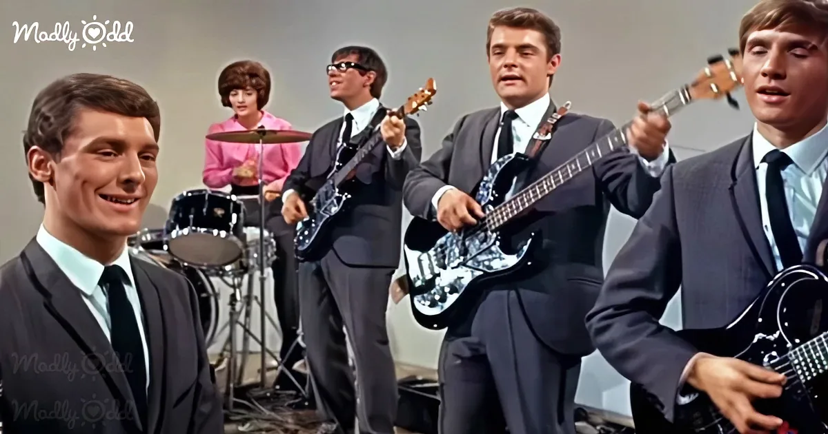 A smiling man in a suit stands in front of a band playing guitars and drums.