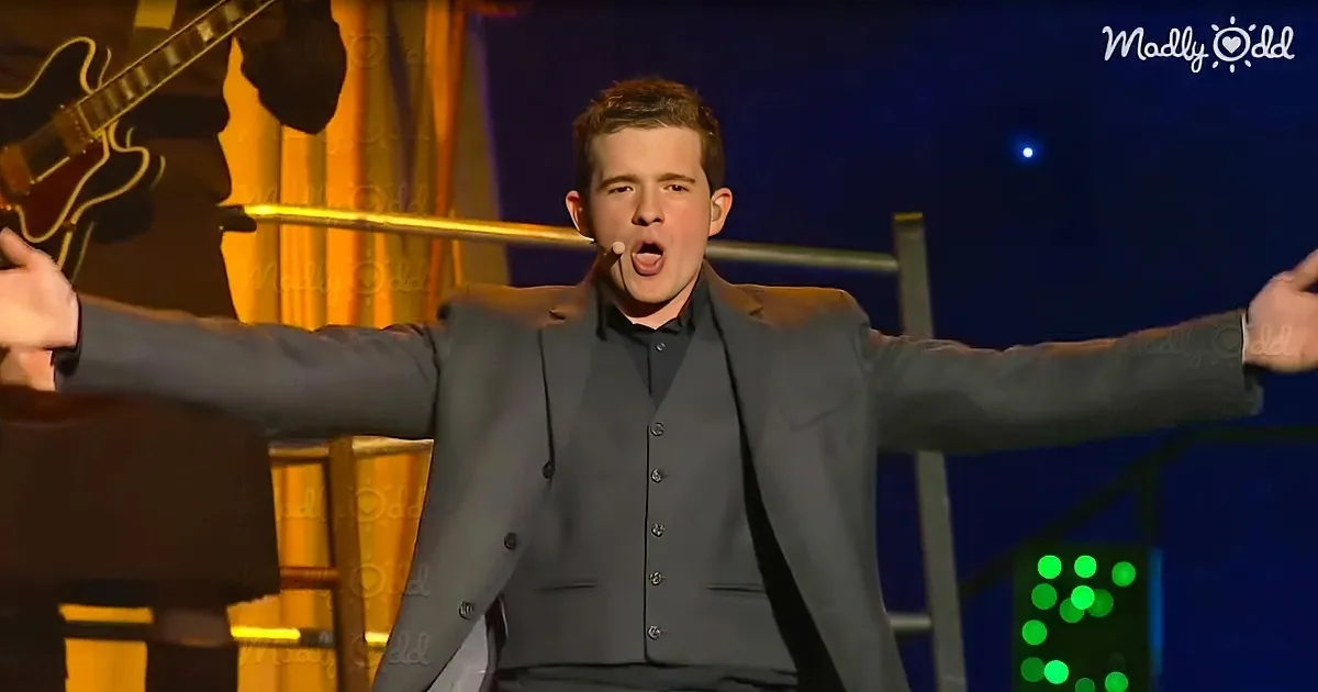 A man in a gray suit sings passionately on stage with arms outstretched, microphone in hand.