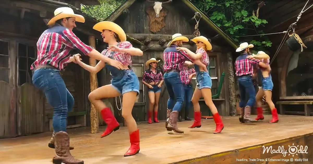 A couple in cowboy attire dances joyfully on a wooden stage, surrounded by other dancers in a rustic setting.