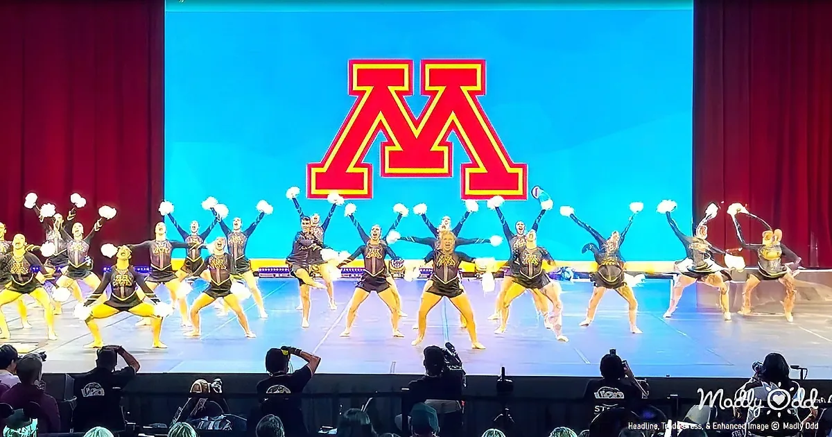 Cheerleaders perform on stage with a large "M" logo in the background.
