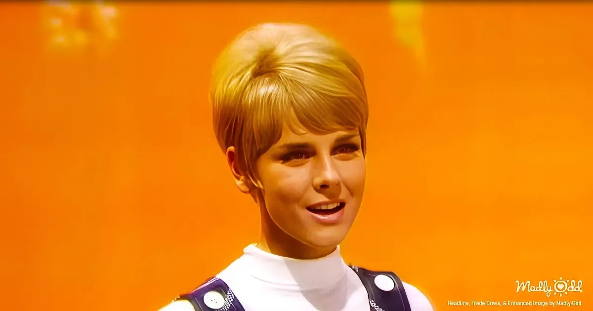 A person with short, light brown hair wearing a white turtleneck and a dark vest with white buttons, standing against an...