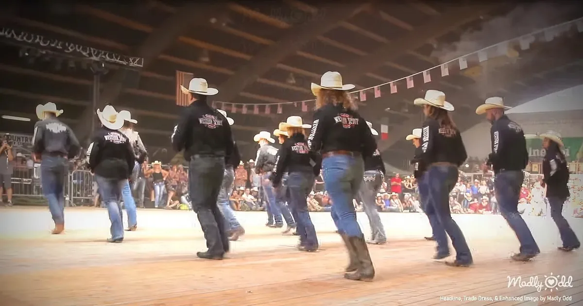 A group of people in cowboy hats and black jackets dances on a wooden stage in front of a crowd.