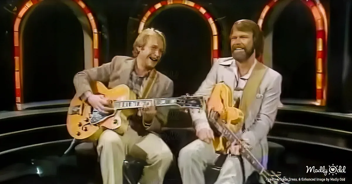 Two men in suits play guitars on a stage with arched, colorful backgrounds.