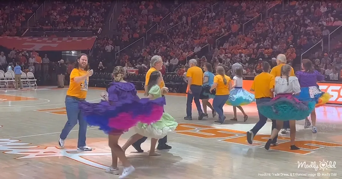 People in colorful skirts dance on a basketball court in front of a large crowd.