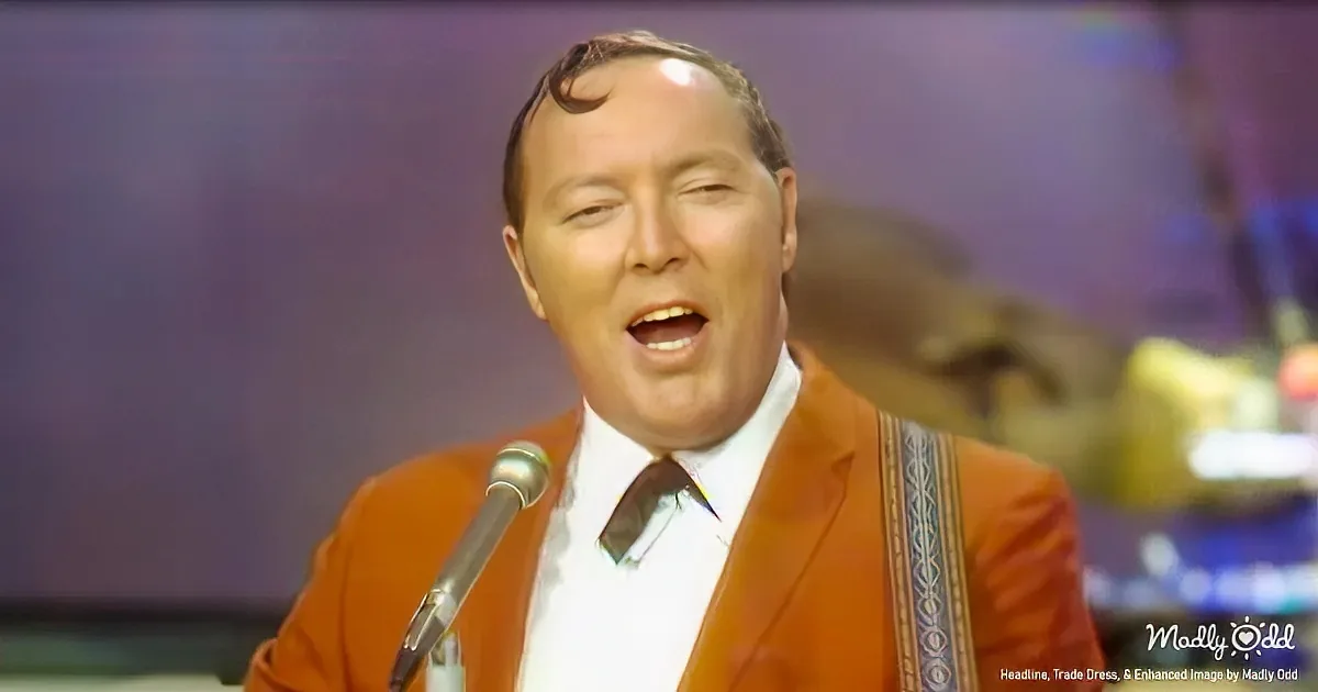 A man in an orange suit sings into a microphone, wearing a white shirt and patterned tie.