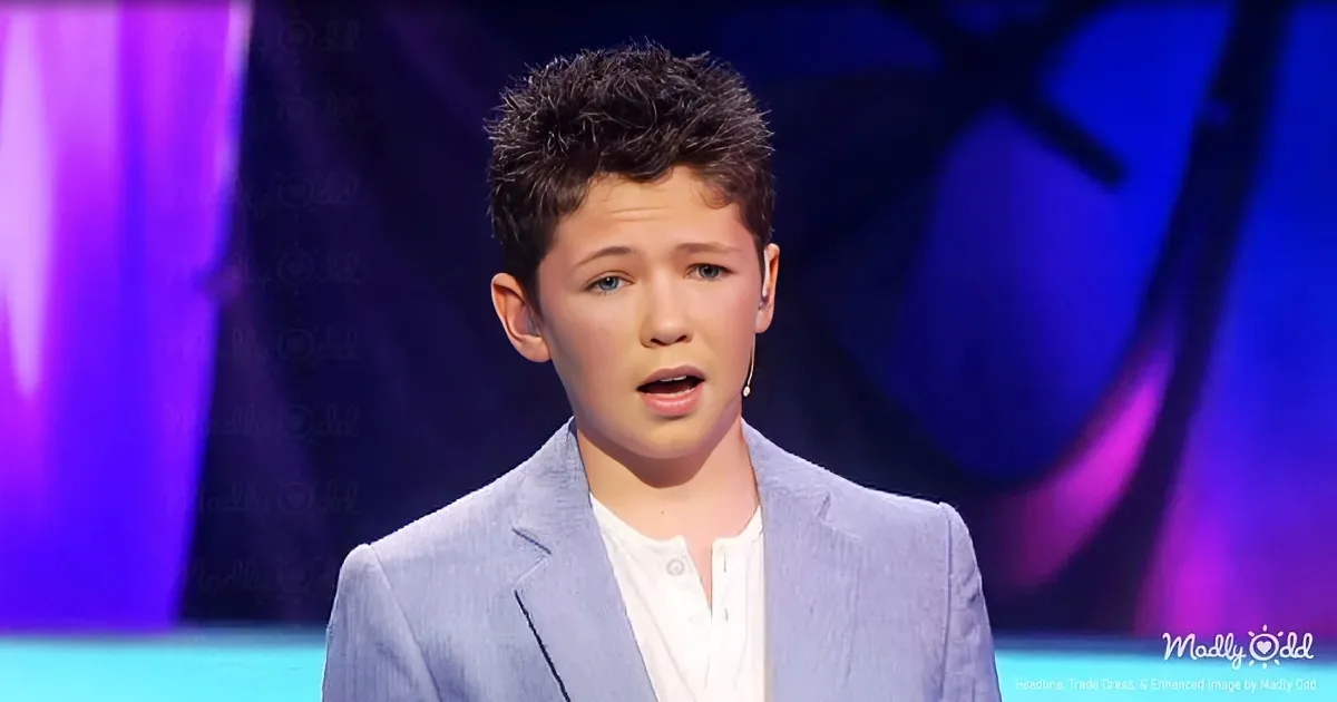 A young person in a light blue suit jacket and white shirt stands on a stage with a purple and blue background.