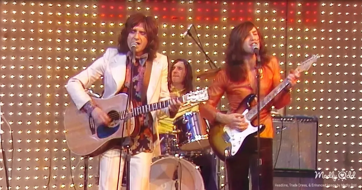 A band performs on stage with a drummer, guitarist, and lead singer, illuminated by a backdrop of lights.