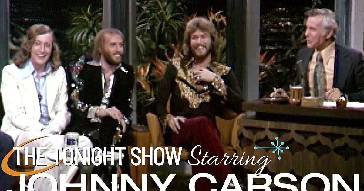 Four men in 70s attire, one in gold, seated, smiling, with drinks on a table.