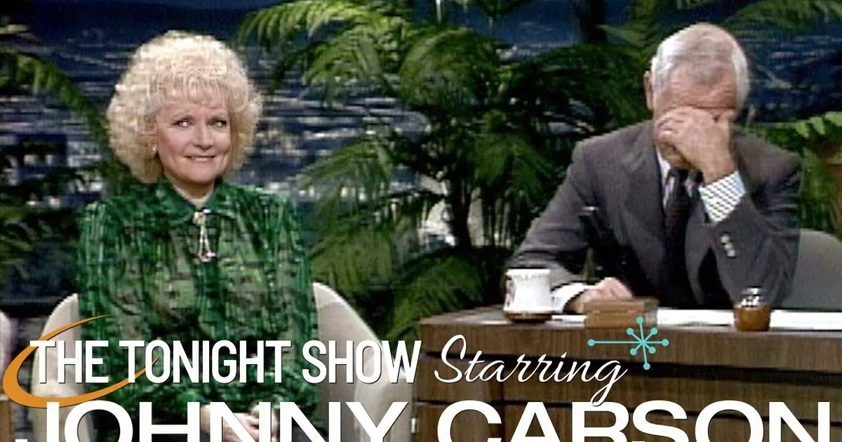 A woman in a green dress laughs while seated next to a man in a suit on a talk show set.