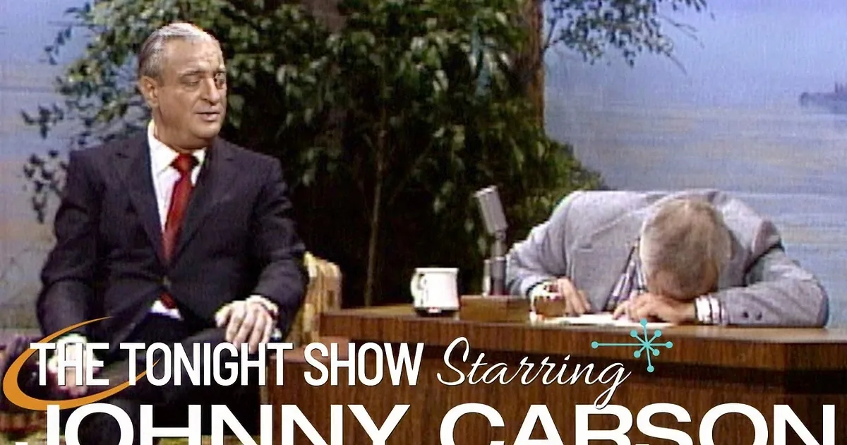 Two men in suits sit at a desk with a microphone, one laughing, the other serious, in a studio setting.